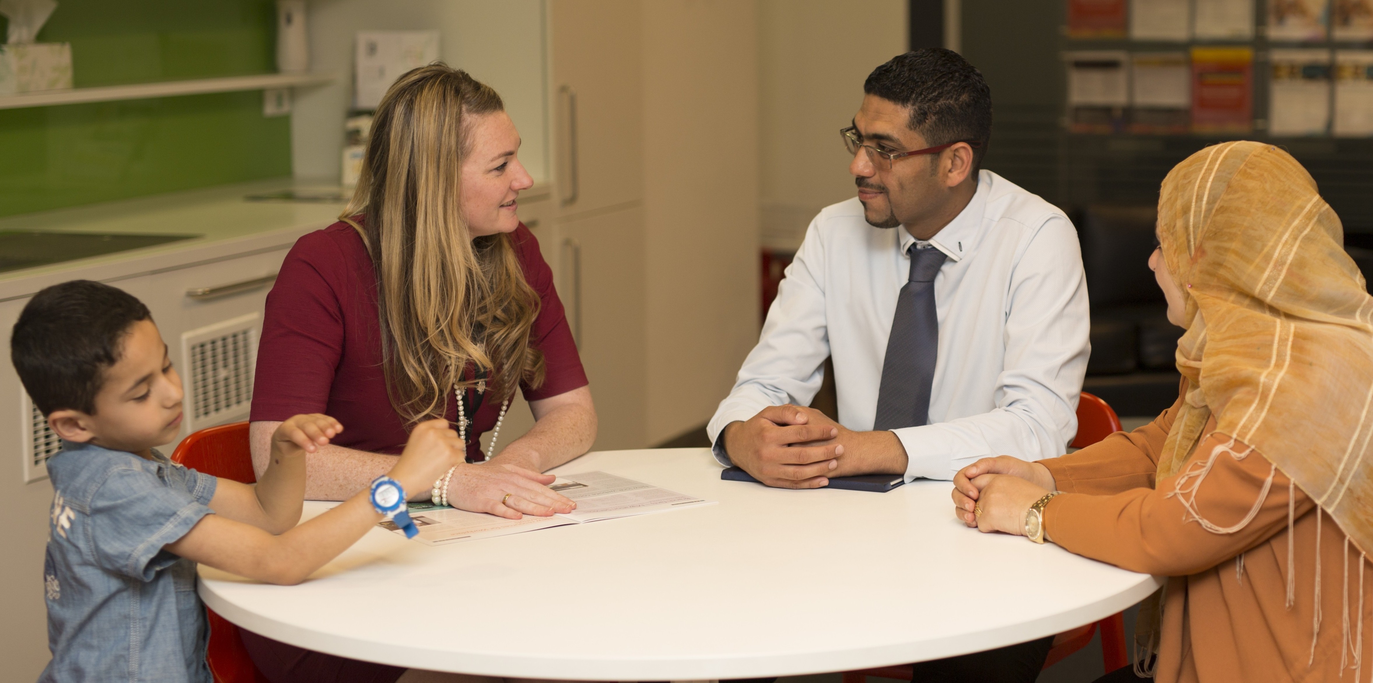 A small boy in a blue shirt, a white woman with blonde hair and a red shirt, a dark skinned man in a white shirt and tie and a woman in an orange top and burqa sit around a table in an office.