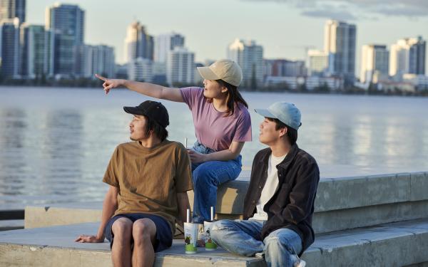 Three young people wearing hats sitting by the river in front of the Perth skyline