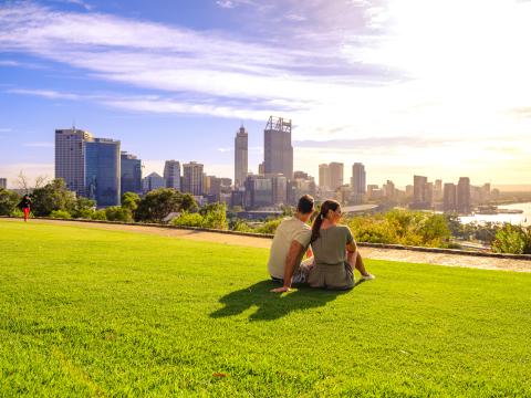 A couple sit on a grassy hill overlooking the Perth skyline. 