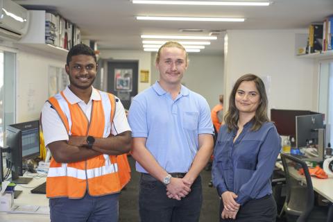 A man in an orange high visibility vest stands in an office next to a man with a blonde moustache in a light blue short sleeved shirt, and a woman with long brown hair and a dark blue button up shirt.