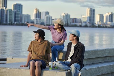 Three young people wearing hats sitting by the river in front of the Perth skyline