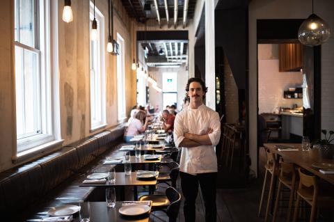 A man wearing a chefs uniform, standing inside a restaurant with his arms crossed.