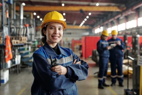 woman skilled migrant standing in a factory with a yellow safety, holding a transceiver and wearing blue jumpsuit