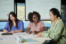 Three women sit at a desk inside an office.