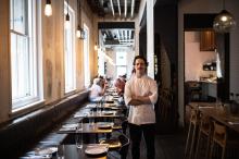 A man wearing a chefs uniform, standing inside a restaurant with his arms crossed.